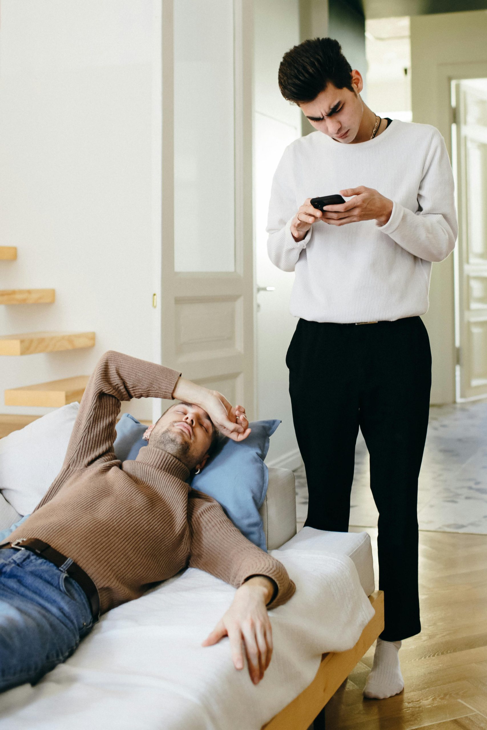 Un jeune homme en pull blanc consulte son téléphone tandis qu'un homme en pull marron se repose sur le canapé, illustrant les dynamiques modernes des relations.