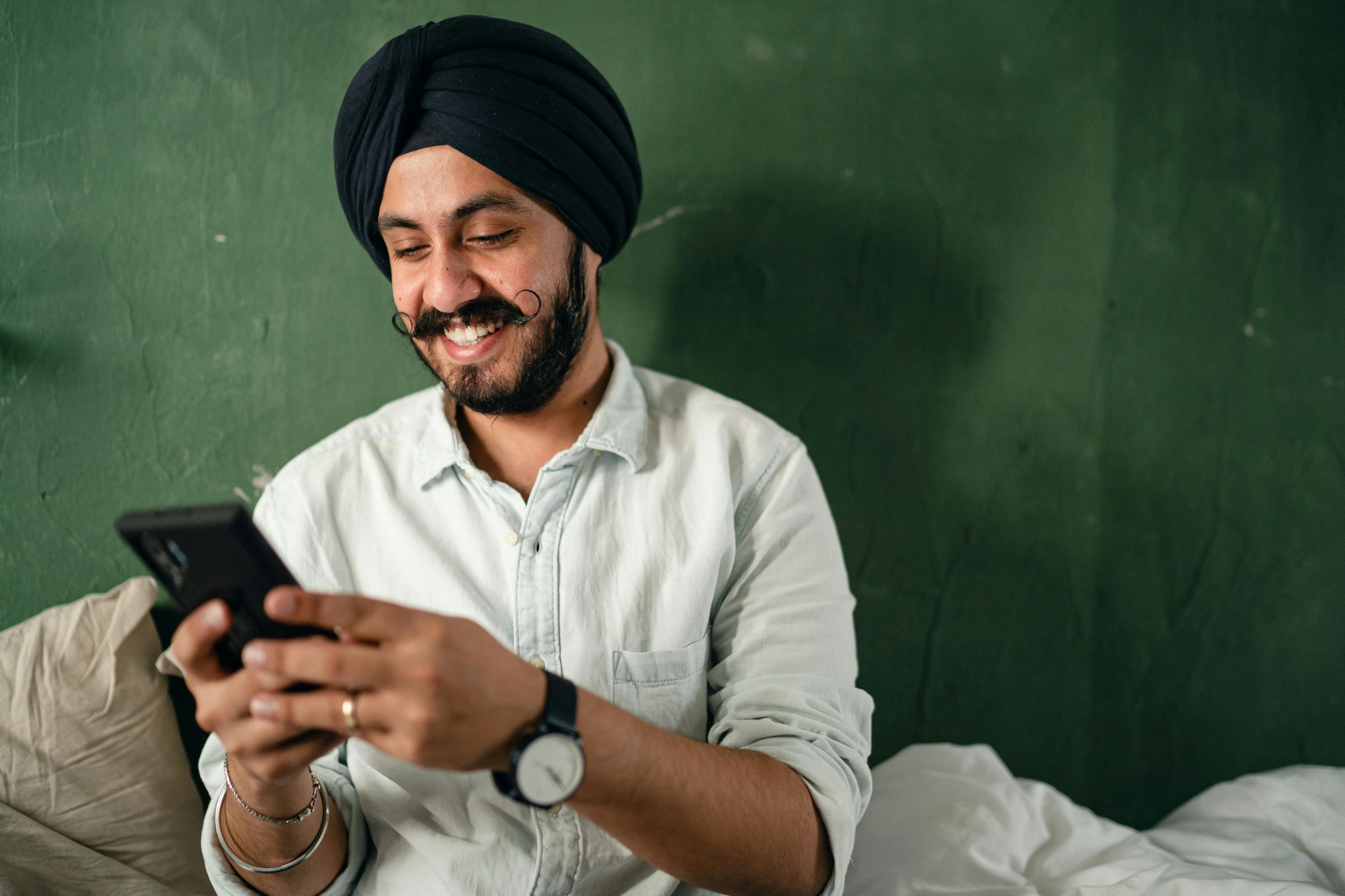 Jeune homme indien avec une barbe et une moustache noires, vêtu d'une chemise décontractée et d'un turban, envoyant des messages sur un smartphone et souriant joyeusement tout en étant assis sur un lit près d'un mur vert à la maison