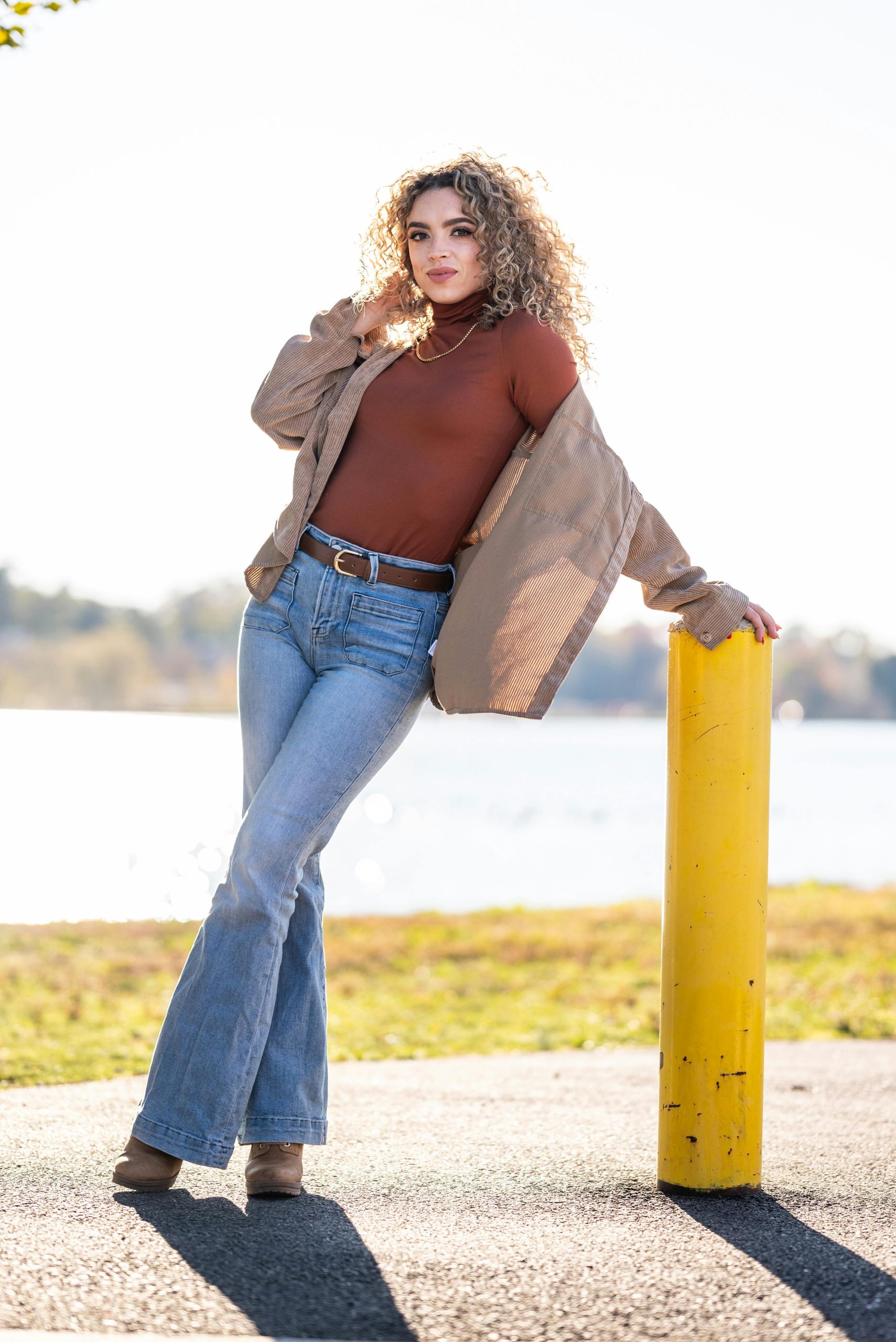 Fashion-forward woman with curly hair posing by a lake on a sunny day in casual attire. Femme à la mode avec des cheveux bouclés posant près d'un lac par une journée ensoleillée en tenue décontractée.