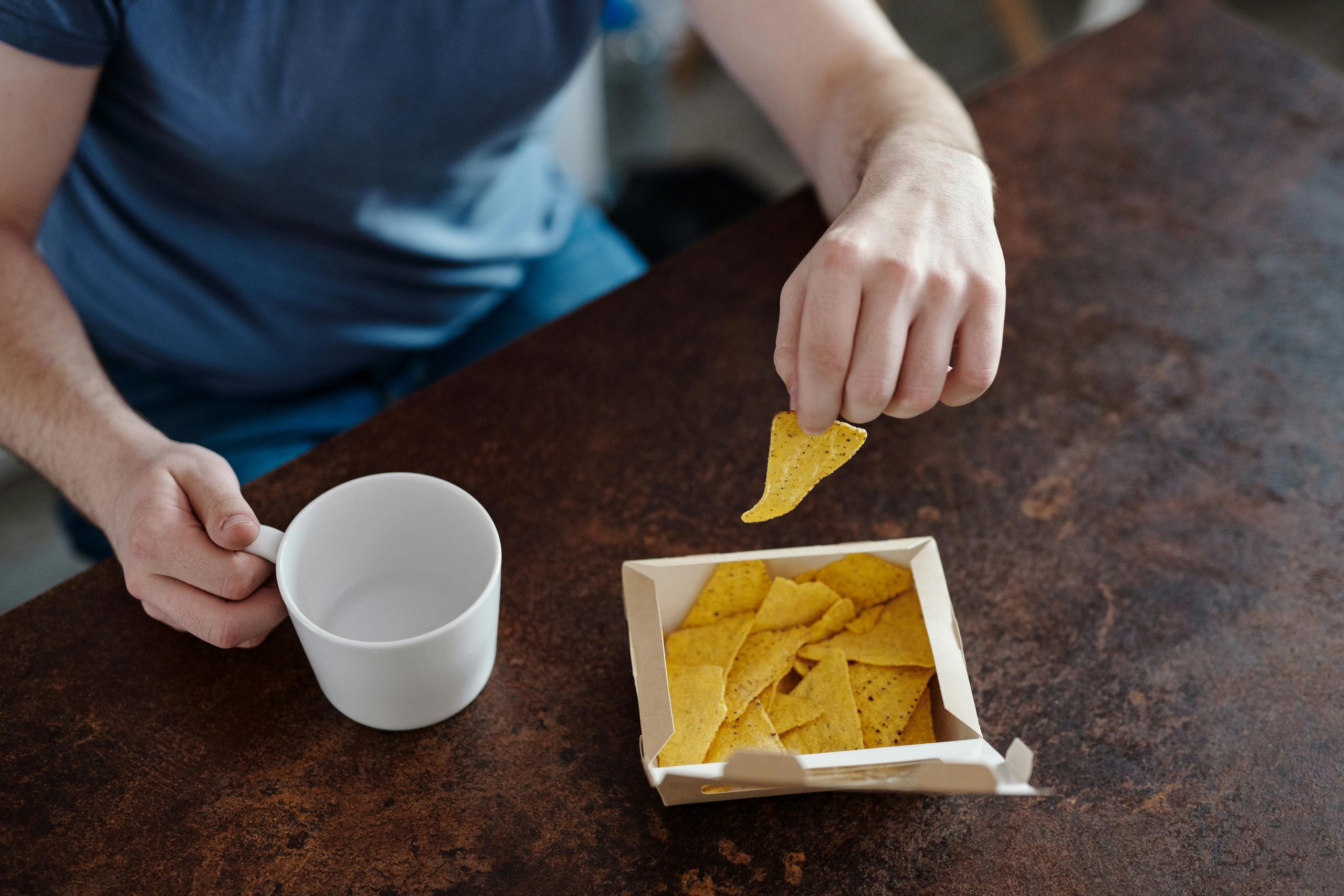 Close-up of a man holding nachos and a ceramic cup, emphasizing casual snack time. Gros plan d'un homme tenant des nachos et une tasse en céramique, soulignant un moment de collation décontracté.