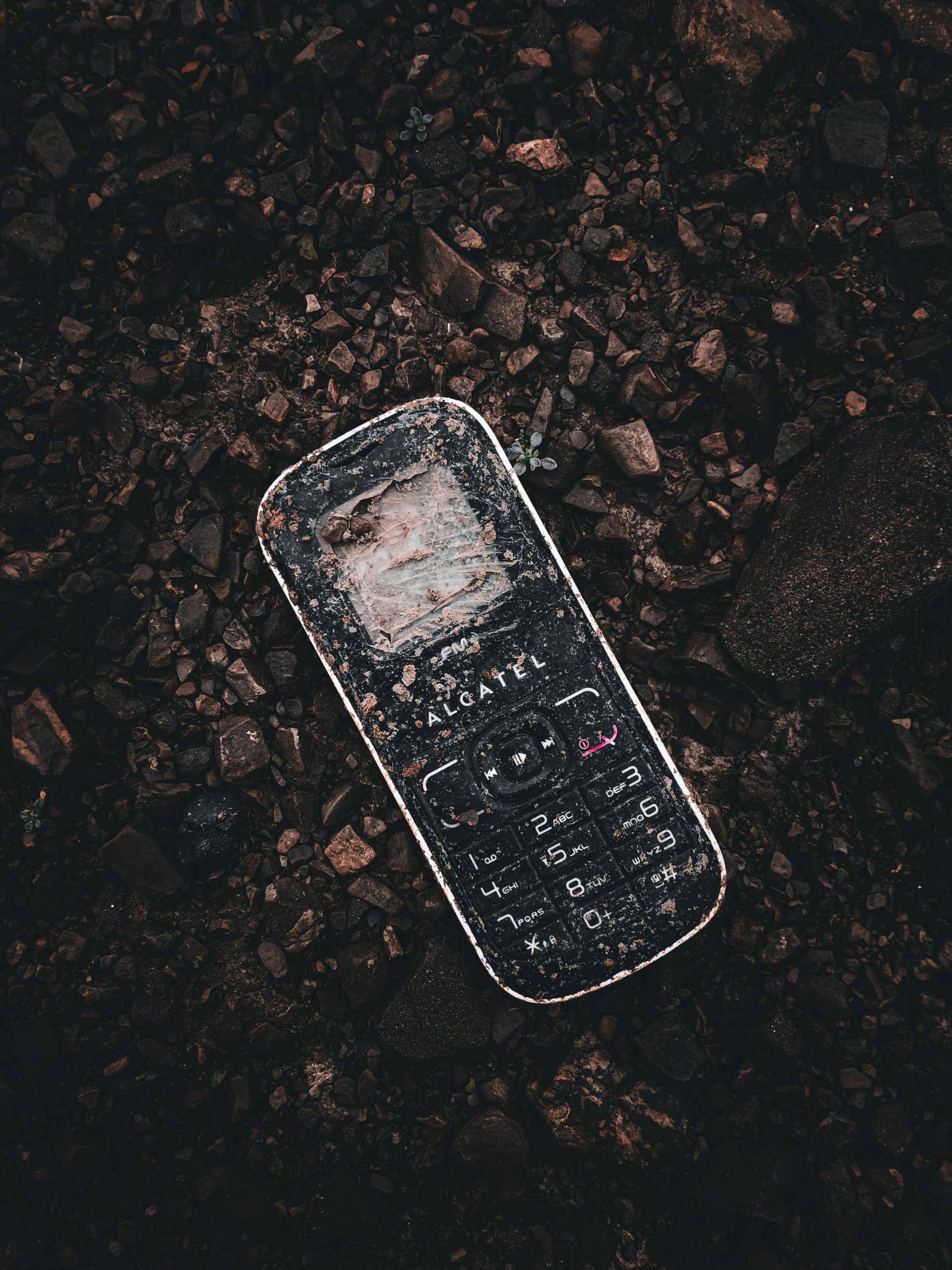 A weathered cellphone lies on rocky ground, showcasing urban decay in Albania. Un téléphone portable usé gît sur un sol rocheux, mettant en avant la décadence urbaine en Albanie.