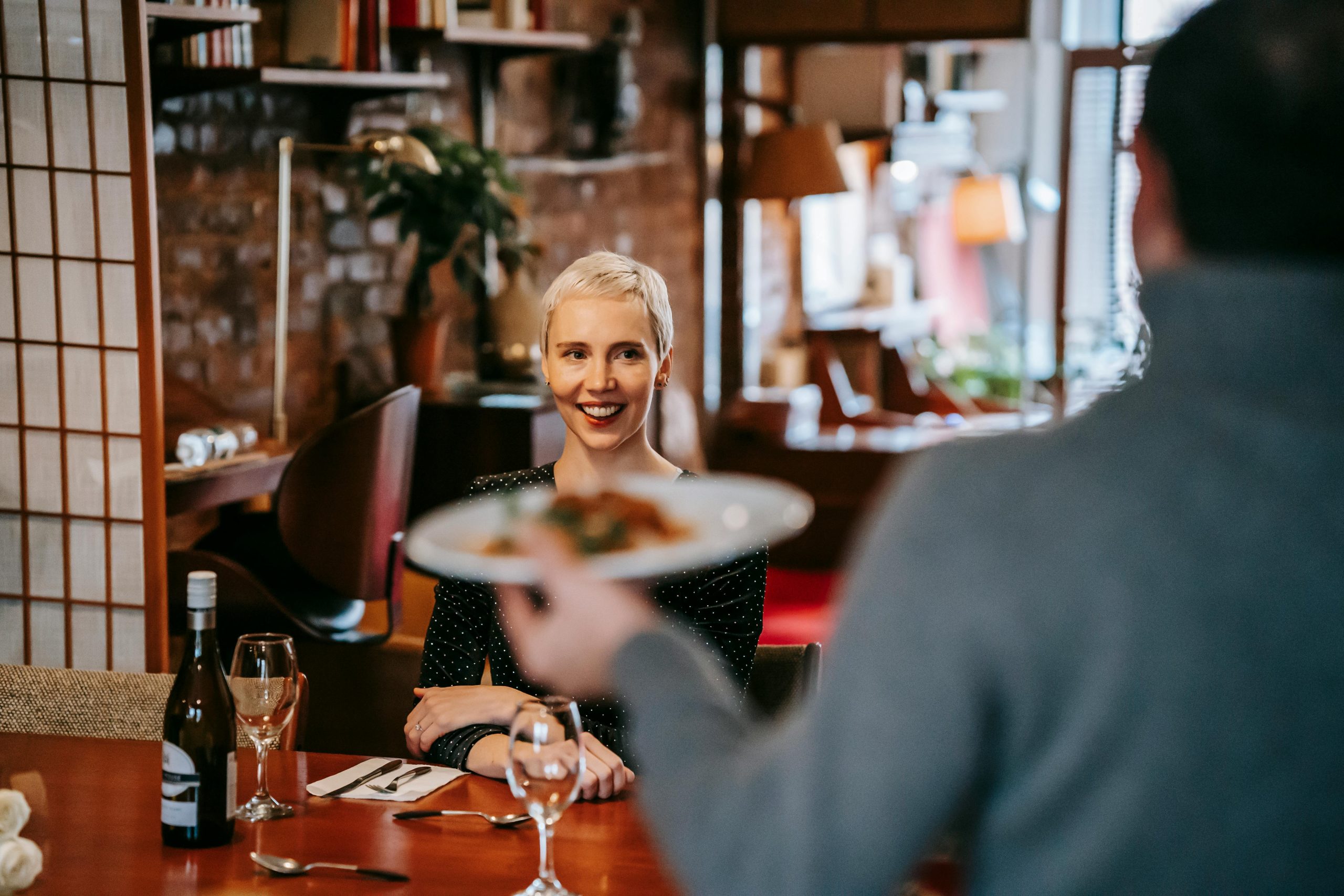Positive couple having date in bright restaurant while man serving plate with food to woman sitting in chair at table with wine bottle and glasses near cutlery with napkin Un couple positif en rendez-vous dans un restaurant lumineux où l'homme sert une assiette de nourriture à la femme assise sur une chaise à table avec une bouteille de vin et des verres près des couverts avec une serviette.