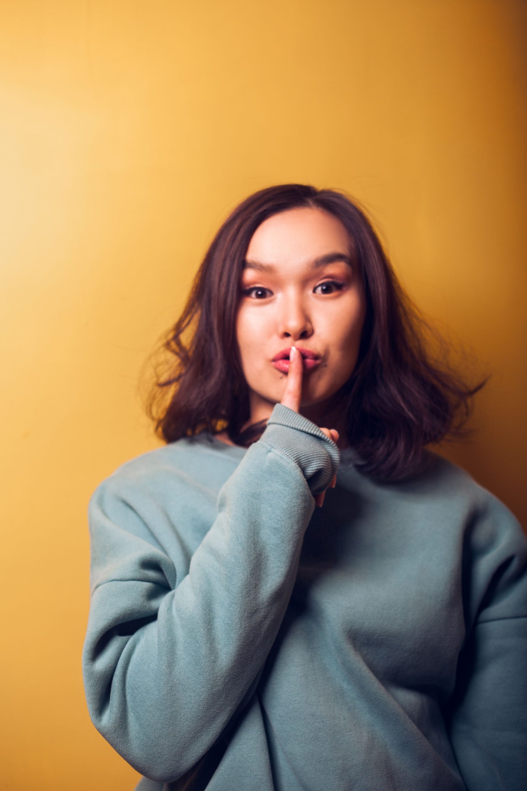Young Asian female in warm clothing doing shh gesture with finger on lips and looking at camera while standing near orange wall Jeune femme asiatique vêtue de vêtements chauds faisant un geste de silence avec le doigt sur les lèvres et regardant la caméra en se tenant près d'un mur orange