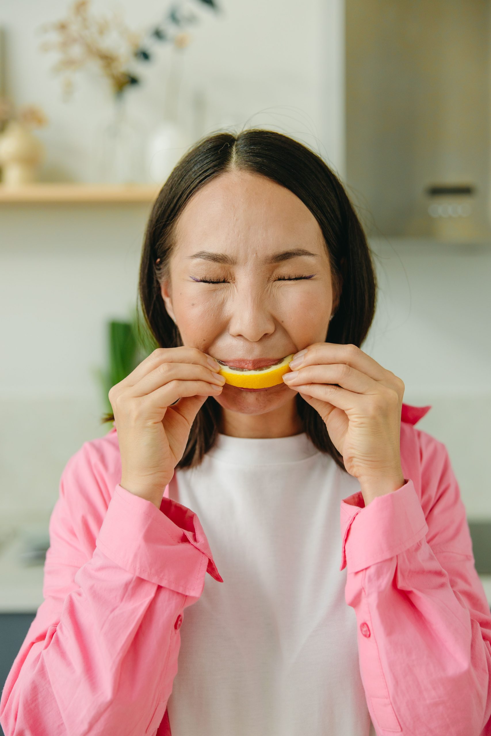 Asian woman in pink shirt makes a funny face while tasting a sour lemon slice indoors. Une femme asiatique en chemise rose fait une grimace amusante en goûtant une tranche de citron aigre à l'intérieur.