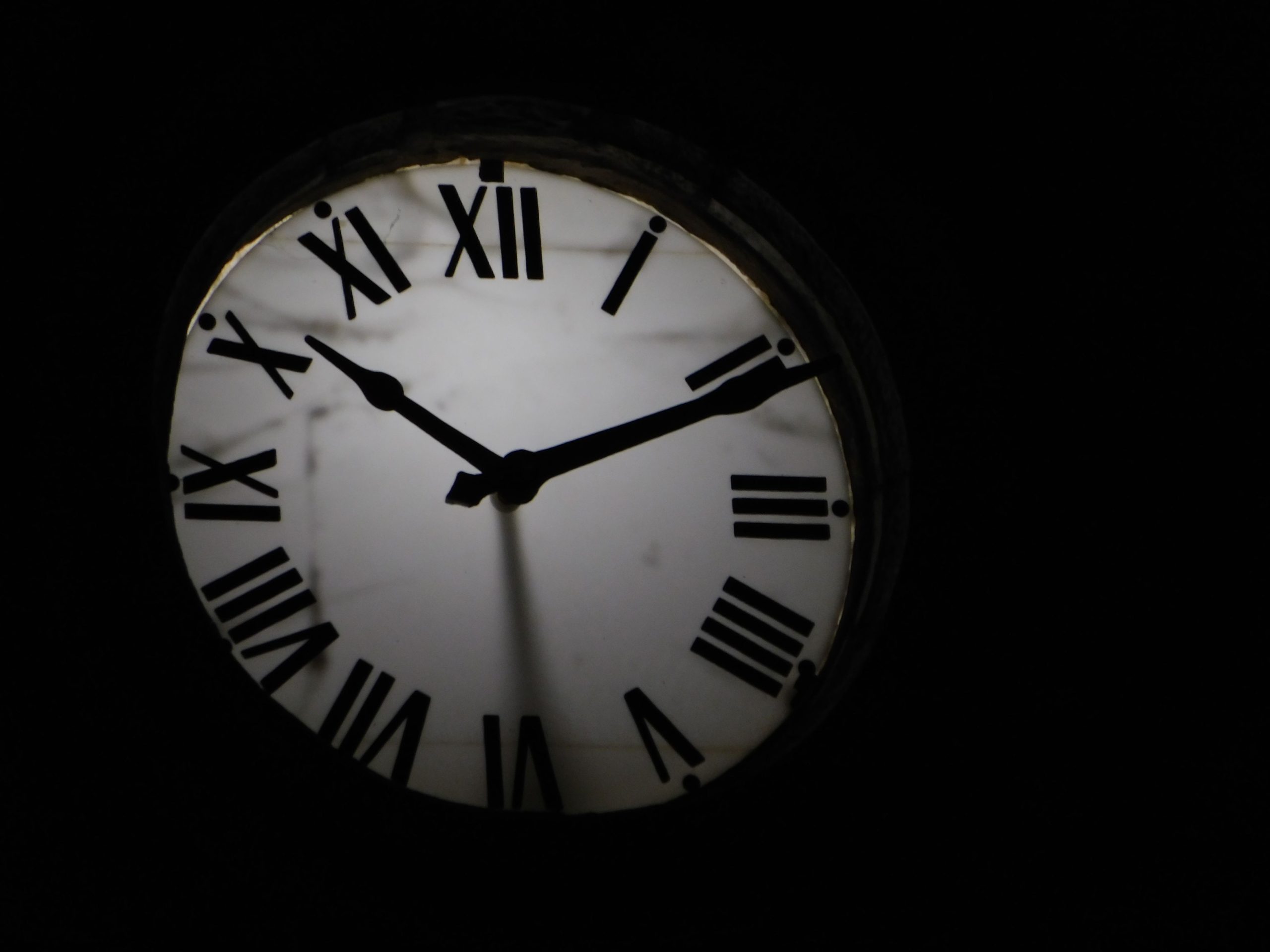 Round mechanical clock with black arrows and numbers on white face ticking on black background in darkness Horloge mécanique ronde avec des flèches noires et des chiffres sur un cadran blanc qui tique sur un fond noir dans l'obscurité