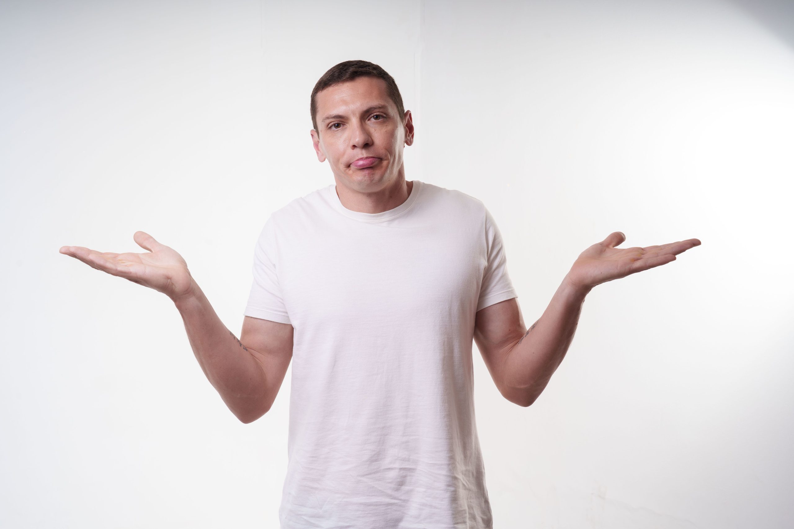 Adult man in white t-shirt shrugs in studio, expressing confusion against a plain background. Homme adulte en t-shirt blanc qui hausse les épaules en studio, exprimant la confusion sur un fond uni.