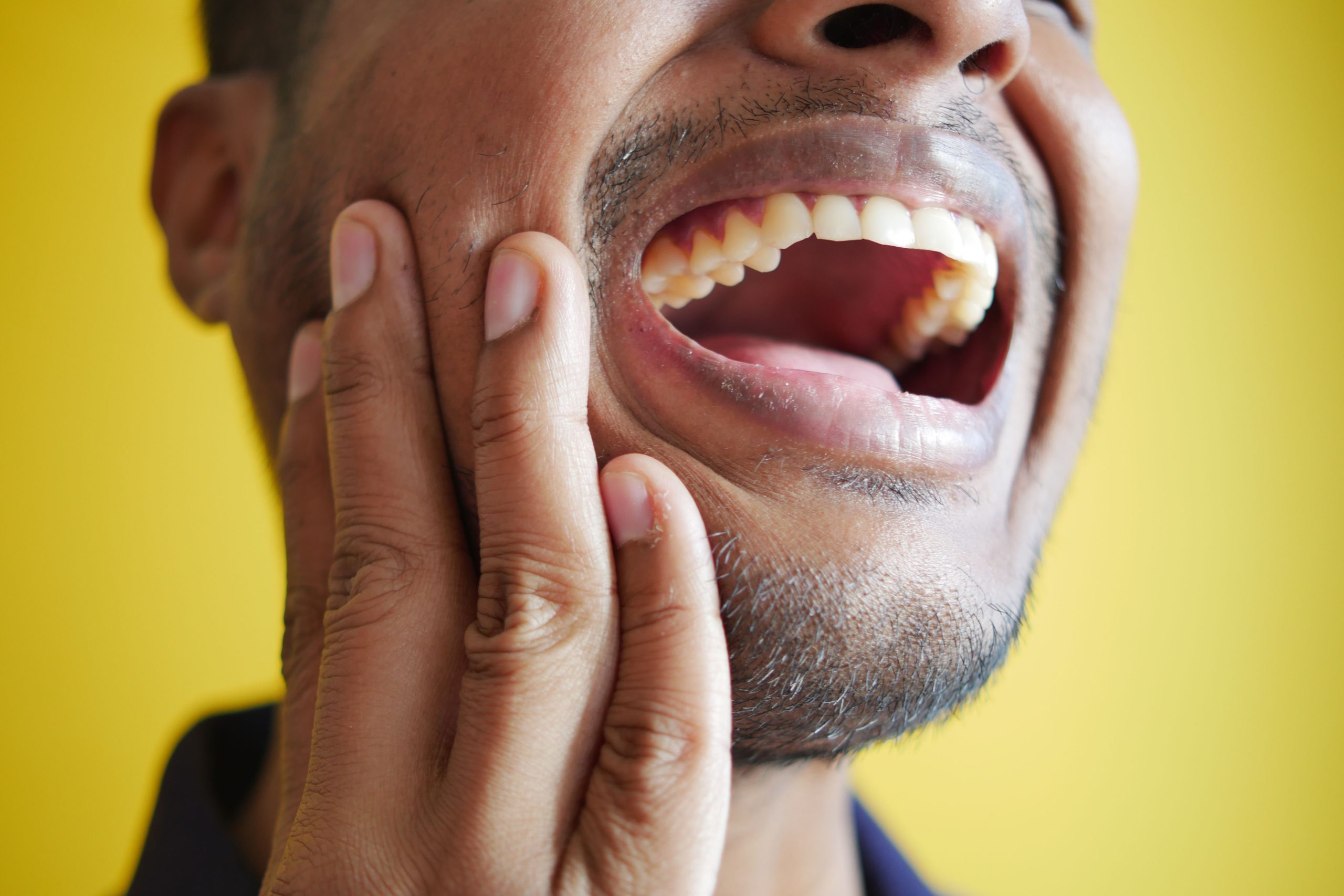 Close-up of a man holding his cheek, displaying a wide open mouth against a yellow background. Gros plan d'un homme se tenant la joue, affichant une bouche grande ouverte sur un fond jaune.