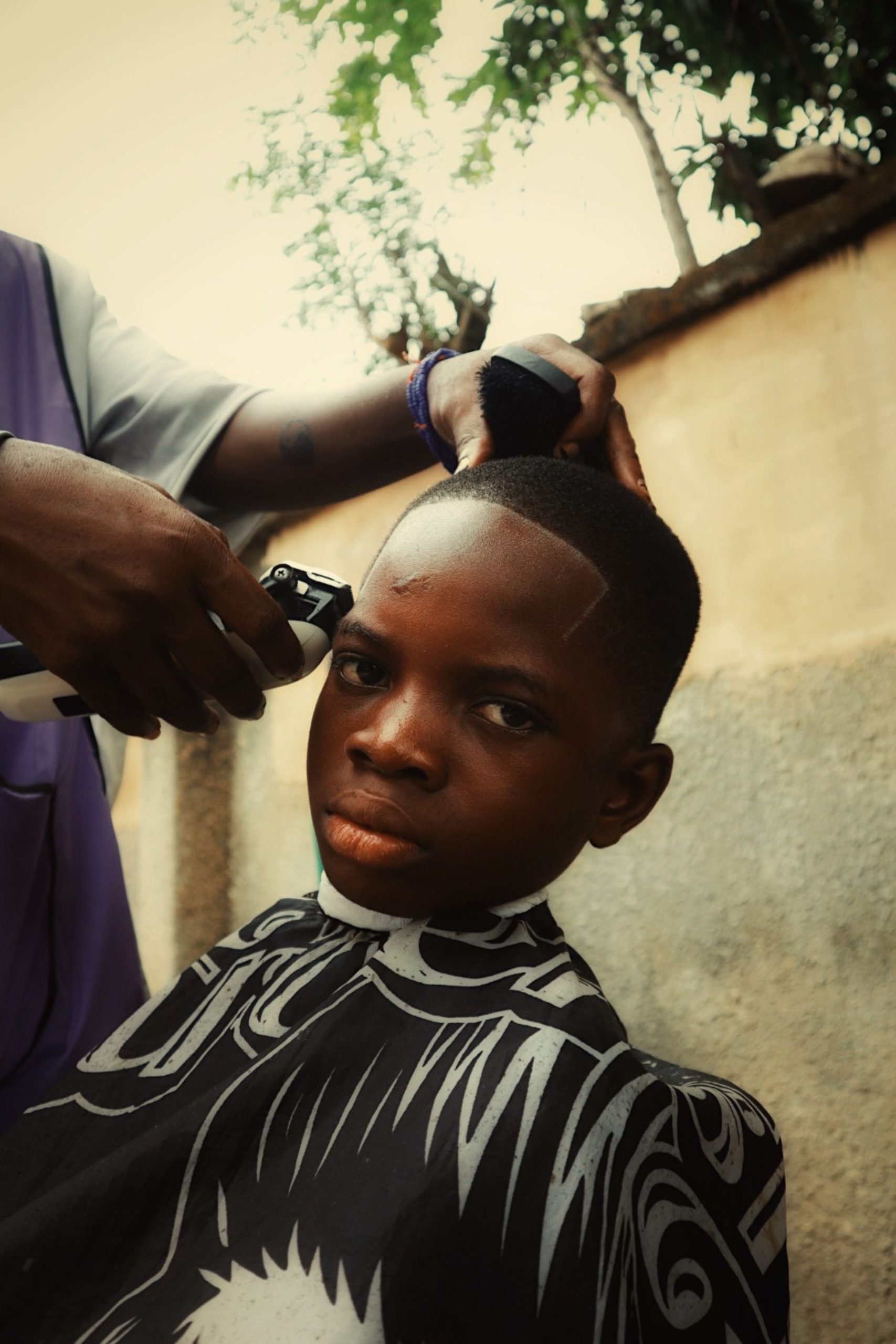 Barber giving a stylish haircut to a young boy outdoors in natural light. Un barbier donnant une coupe de cheveux élégante à un jeune garçon en plein air à la lumière naturelle.