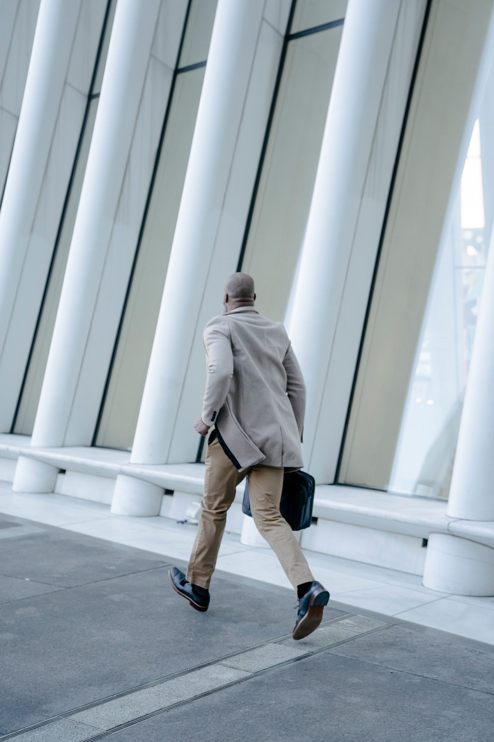 Back view of a man in a brown suit running with a briefcase along architectural columns. Vue arrière d'un homme en costume marron courant avec une serviette le long de colonnes architecturales.