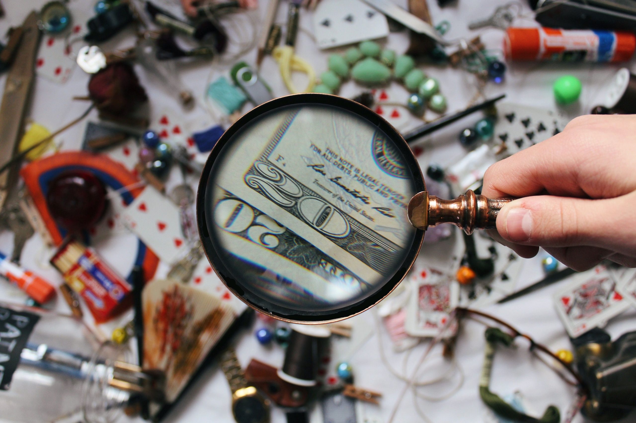 A hand holding a magnifying glass focusing on a twenty dollar bill amidst various scattered objects. Une main tenant une loupe se concentrant sur un billet de vingt dollars parmi divers objets éparpillés.