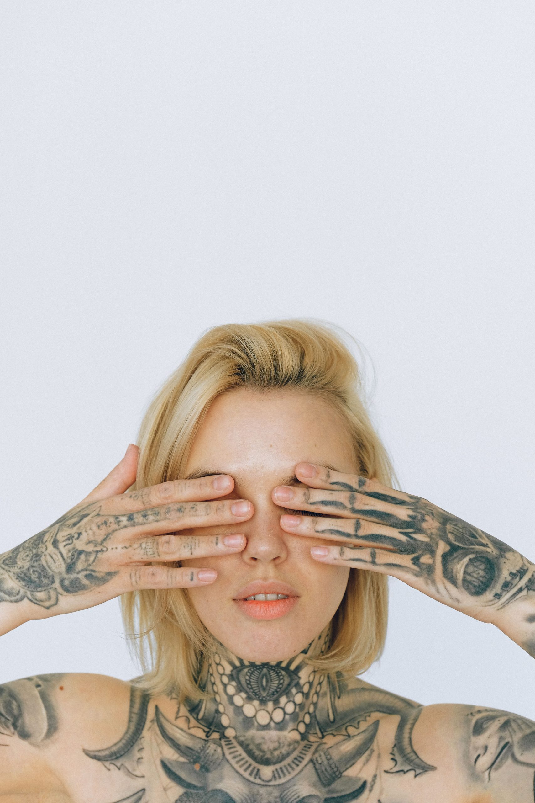 Blonde woman with elaborate tattoos covering her eyes with hands against a white background. Femme blonde aux tatouages élaborés se couvrant les yeux avec les mains sur fond blanc.
