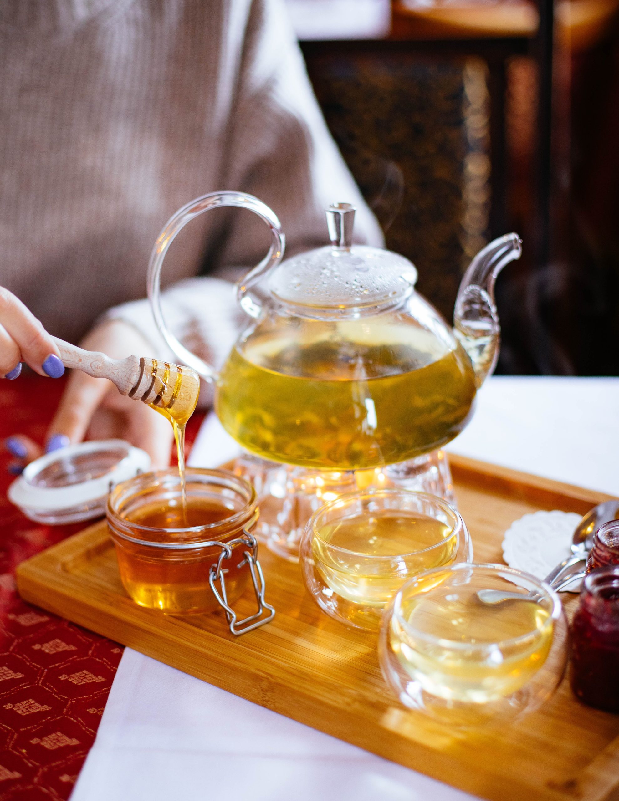 A cozy scene of honey being added to steaming herbal tea in a glass teapot, perfect for relaxation. Une scène douillette montrant du miel versé dans un thé aux herbes fumant dans une théière en verre, parfait pour se détendre.